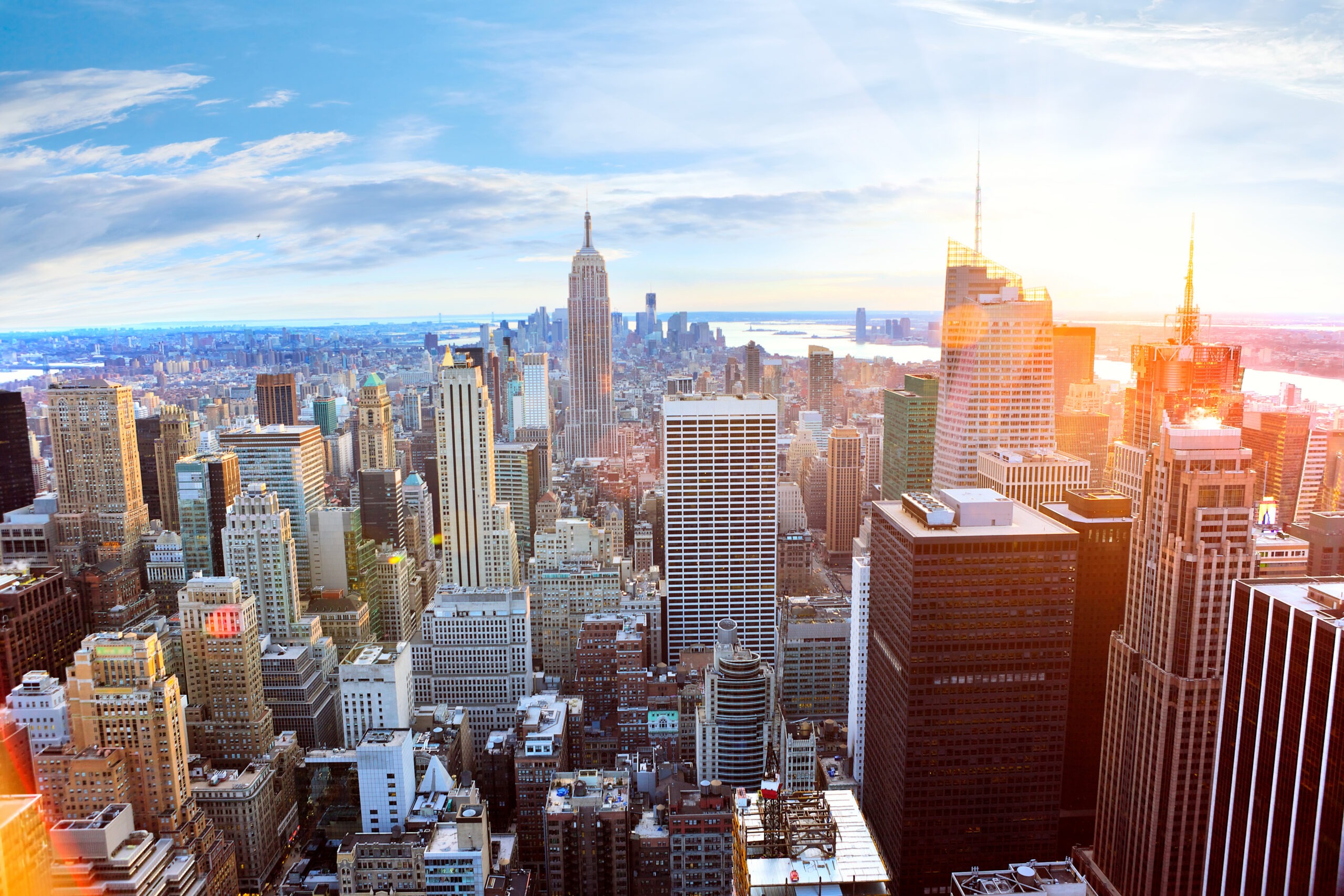 Panoramic view of New York City skyline at sunset, featuring the Empire State Building among modern skyscrapers and the Hudson River in the distance.
