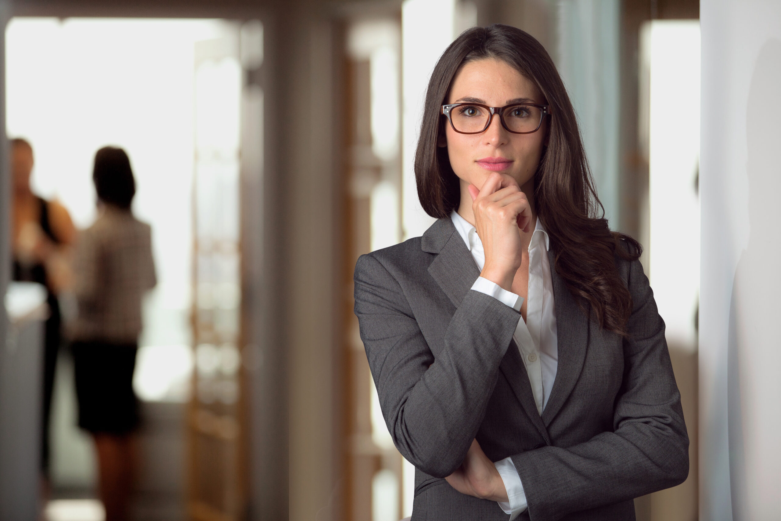 A confident woman in a gray suit stands thoughtfully with her hand on her chin, contemplating, while blurred colleagues converse in the background.